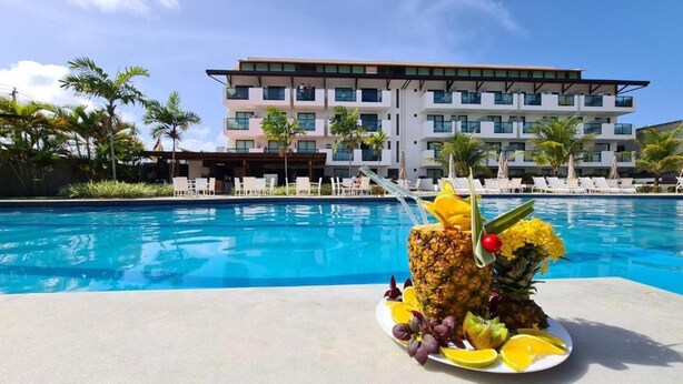 A poolside view with a tropical fruit drink on a plate, palm trees, and a hotel building in the background. - Laguna Beach - Charming, furnished, and cozy loft-style apartment - 402 (Ipojuca)