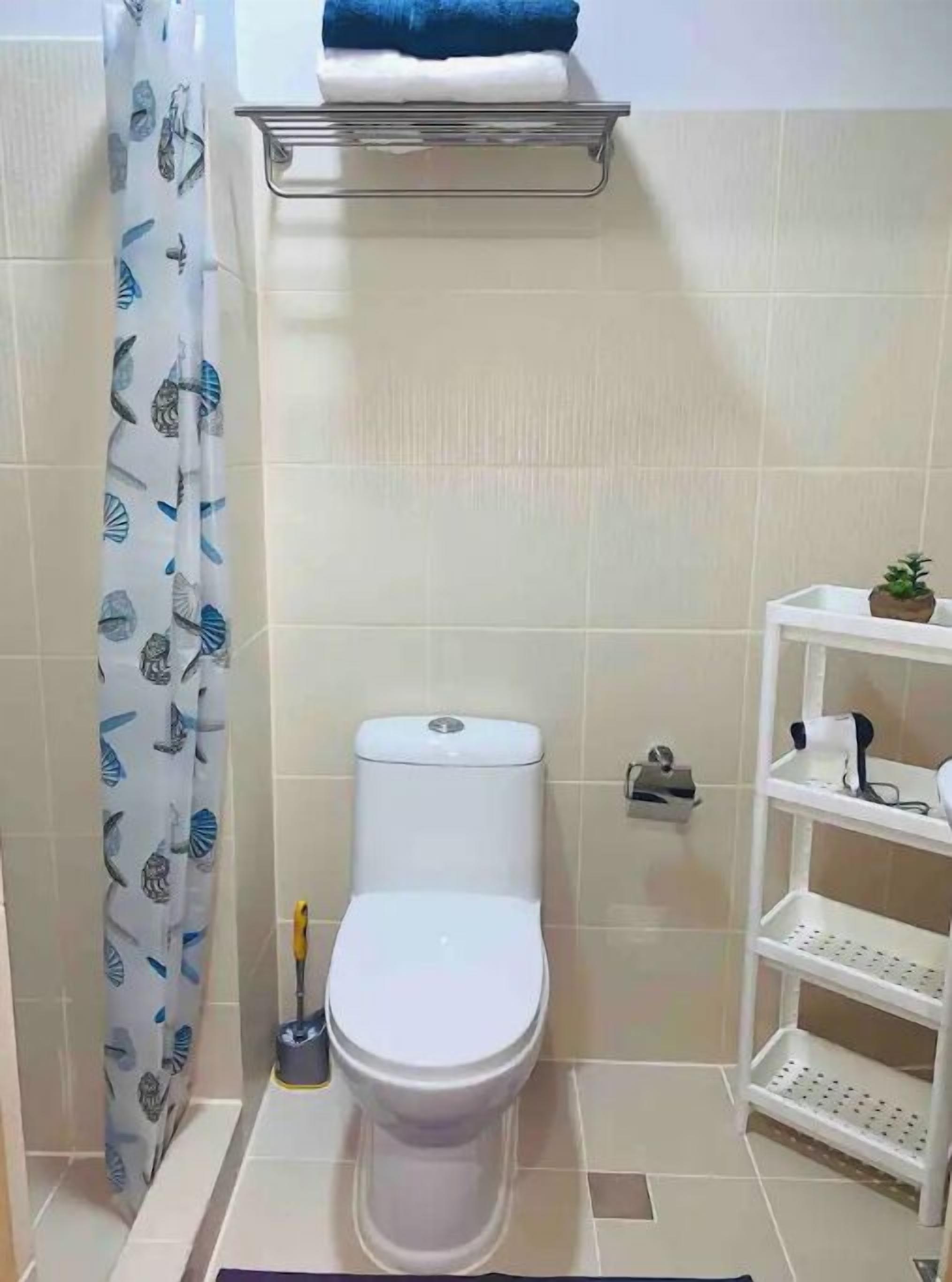 Une salle de bain comprenant des toilettes, une étagère au-dessus des toilettes, une petite étagère sur le côté droit et un rideau de douche orné d’un motif de feuilles.