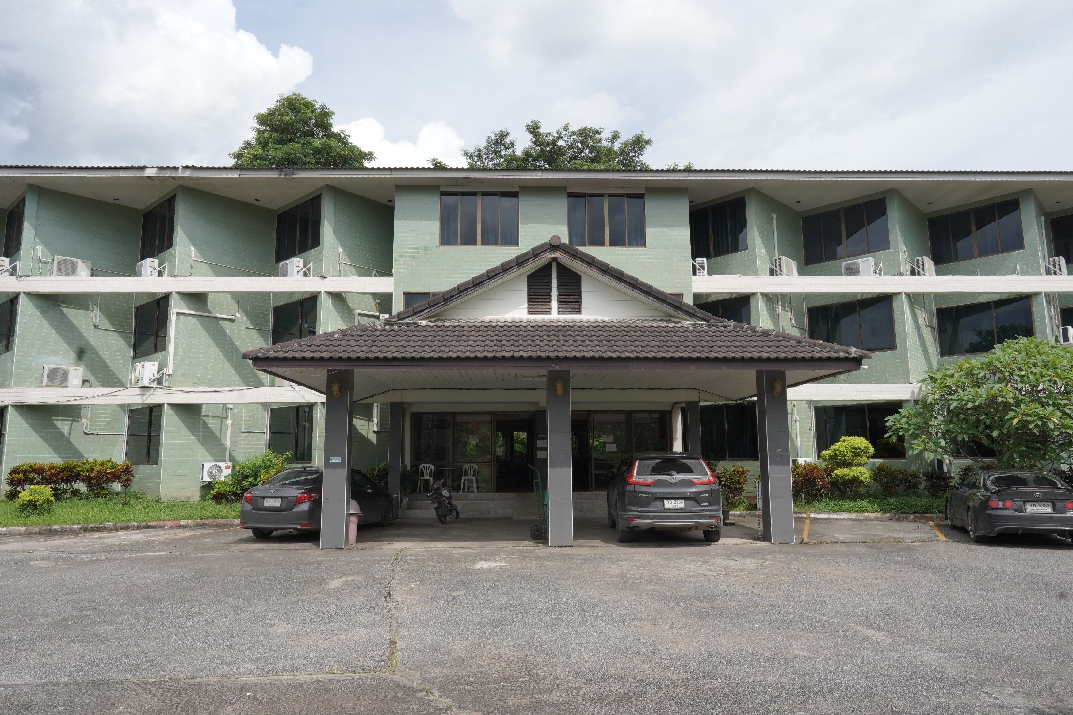 A two-story building with a covered entrance, air conditioning units, and a parking area with cars.