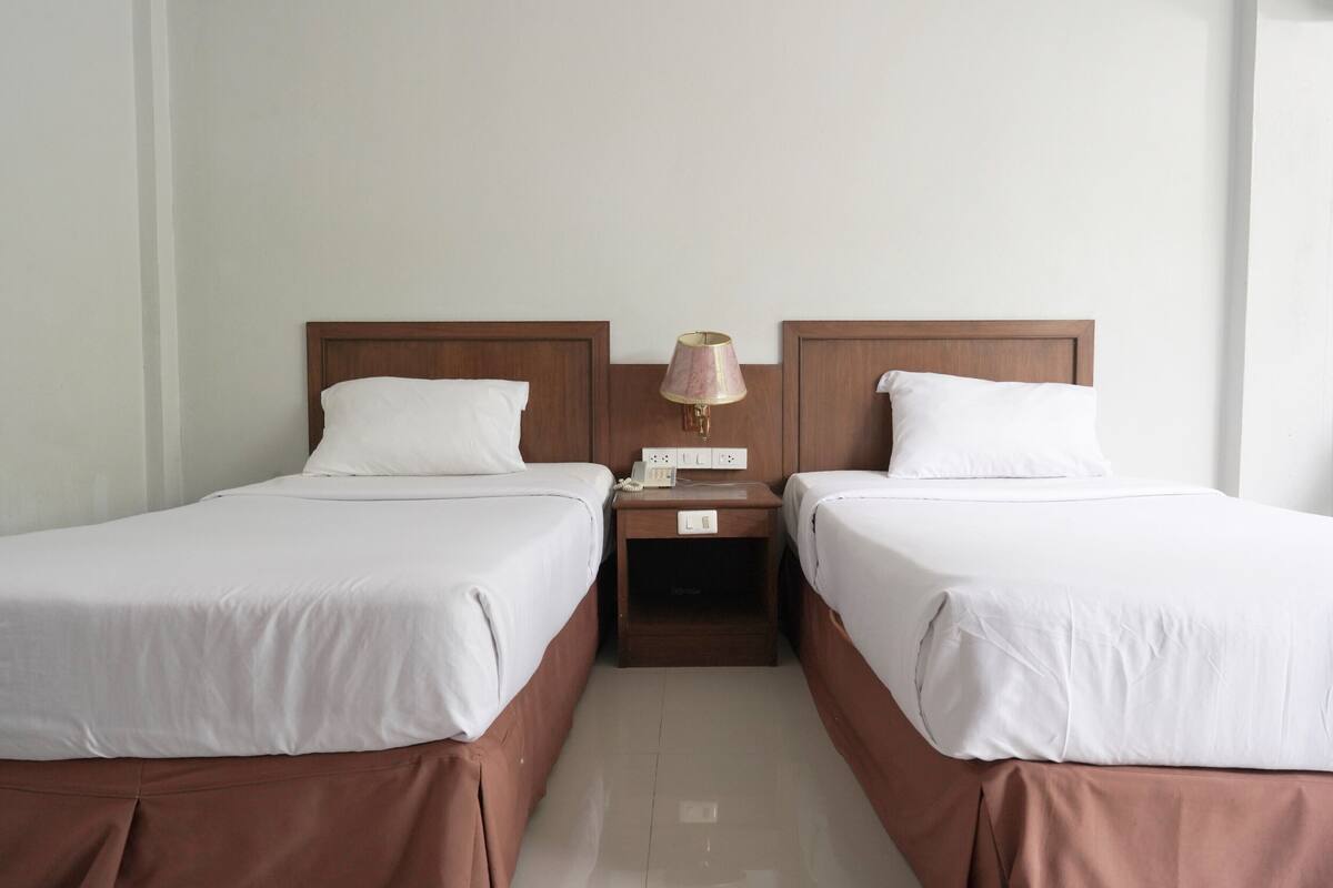 A hotel room with two single beds, a nightstand with a lamp, and a power outlet.