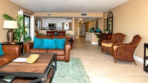 A spacious living room with a brown leather sofa, teal cushions, and a wooden coffee table. A dining area with a wooden table and chairs is visible in the background.