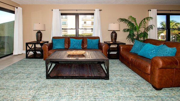 A living room with a brown leather sofa, a wooden coffee table, and two side tables with lamps. There is a large window with white curtains, and a potted plant is visible. - The Villas A-5 - Premium Plus 3 Bed/2 Bath Oceanfront Condo (Clearwater)