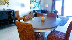 A modern living room with a round dining table, wicker chairs, a ceiling fan, and a large window with curtains.