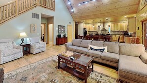 A spacious living area with a high ceiling, wooden staircase, and a kitchen in the background. - Claralea Lodge by Taylor Made Deep Creek (Oakland)