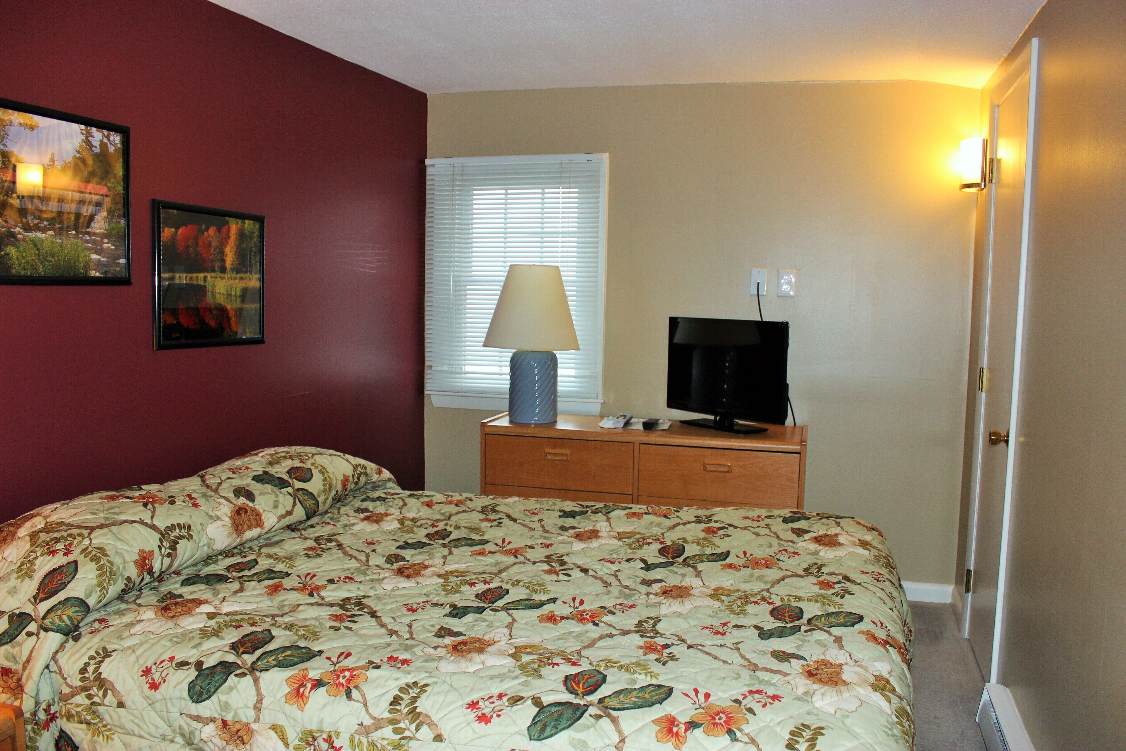 A bedroom with a floral bedspread, a wooden dresser, a television, and two framed pictures on the wall.
