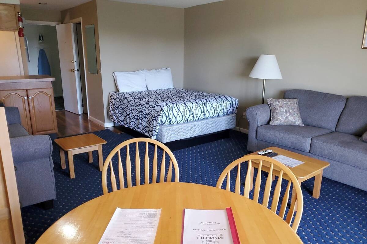 Ein Hotelzimmer mit einem Bett, einem Sofa, einem Schreibtisch und einem Sessel. Auf dem Schreibtisch liegen Papiere und auf dem Sofa steht eine Lampe.