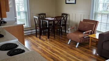 A living room with a wooden floor, a dining table with chairs, a sofa, and a kitchen area with a stove.