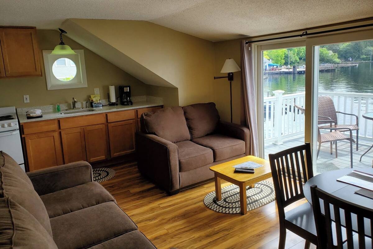 Ein gemütlicher Wohnbereich mit einer Couch, einem Couchtisch und einem Esstisch mit Stühlen. Es gibt eine Küche mit Spüle und Herd. Ein großes Fenster bietet Blick auf ein Gewässer und einen Steg.