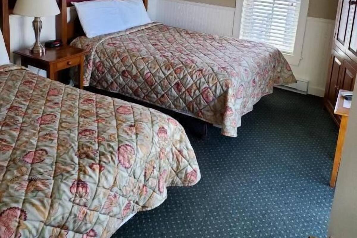 Ein Hotelzimmer mit zwei Betten, einem Nachttisch mit Lampe, einem Fenster mit Jalousien und einer Tür mit Messinggriffen.