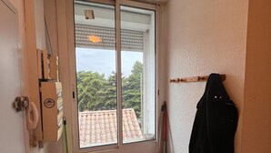 Un baño pequeño con ventana, un trapeador verde, una escoba, una chaqueta negra colgada en la pared y un cubo de basura.