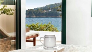 A room with a view of a body of water and hills, a white mug on a coaster, and a book on a table. - Ideal location close to the beach (Skiathos)