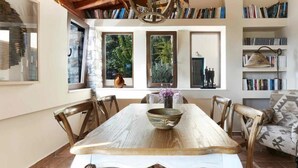 A dining area with a wooden table, chairs, and a chandelier. There are bookshelves and a window with a view of greenery. - Ideal location close to the beach (Skiathos)