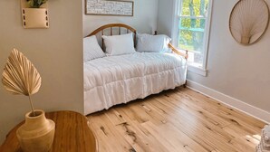 Un dormitorio con piso de madera, una cama con ropa de cama blanca, una lámpara de pared, una ventana con vista a árboles y una planta en la pared.