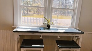 Un escritorio de madera con dos taburetes negros, una planta en maceta y vista a un parque a través de una ventana con marcos blancos.