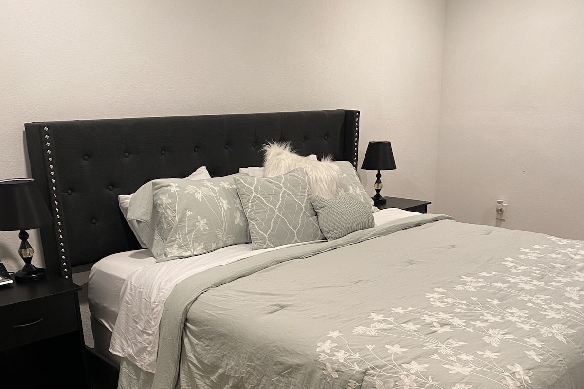 A bedroom with a large bed, a nightstand with a lamp, and a wall socket.