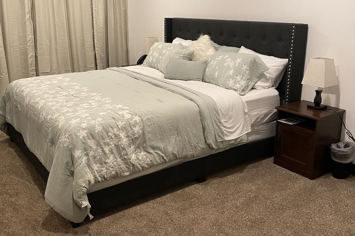A bedroom with a bed, a nightstand, a lamp, and a trash bin.