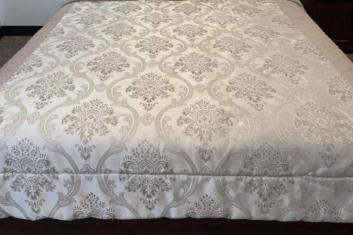 A neatly made bed with a patterned bedspread and several pillows, a bedside table with a lamp and phone, and a plain wall in the background.