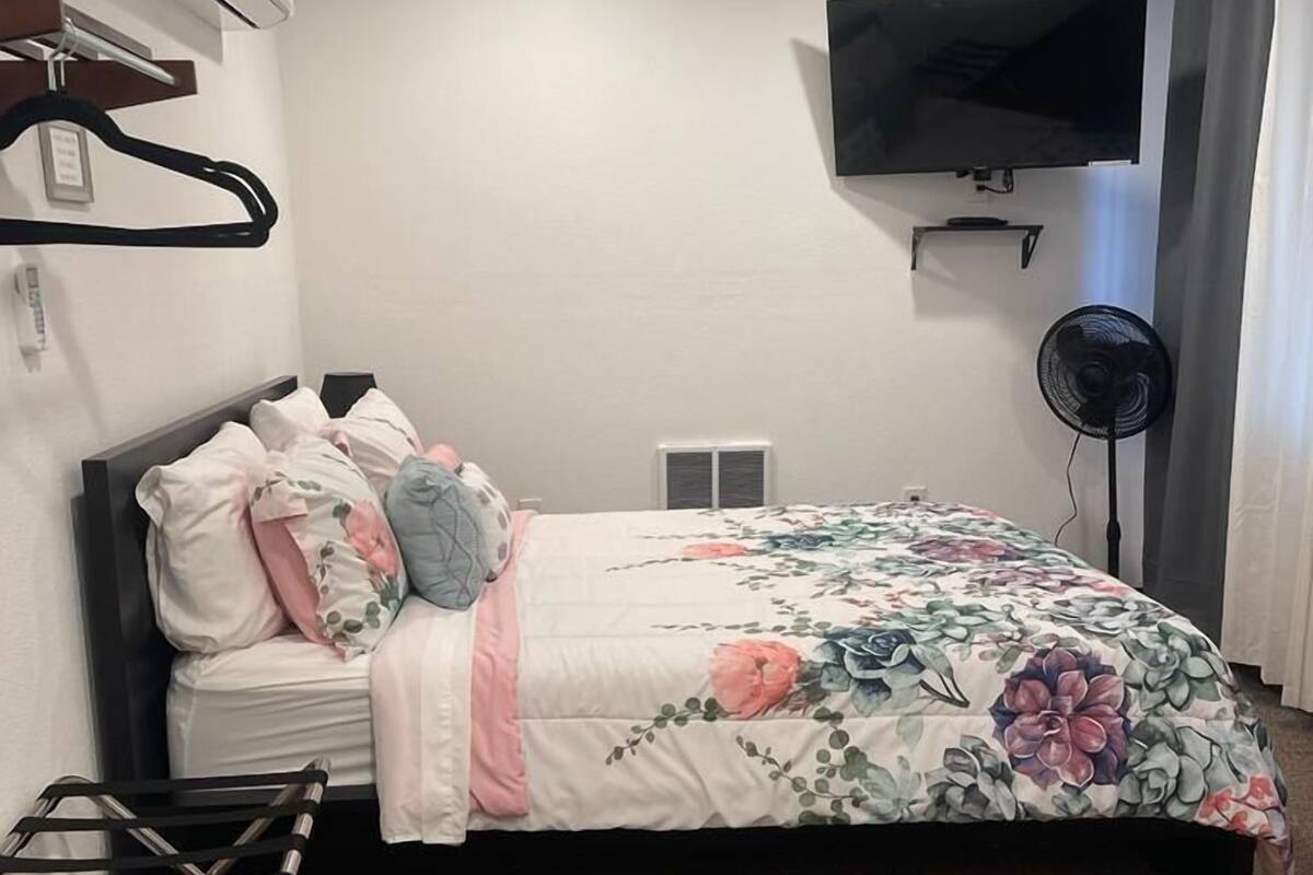 A bedroom with a bed, a TV mounted on the wall, a fan, and a hanging clothes rack.