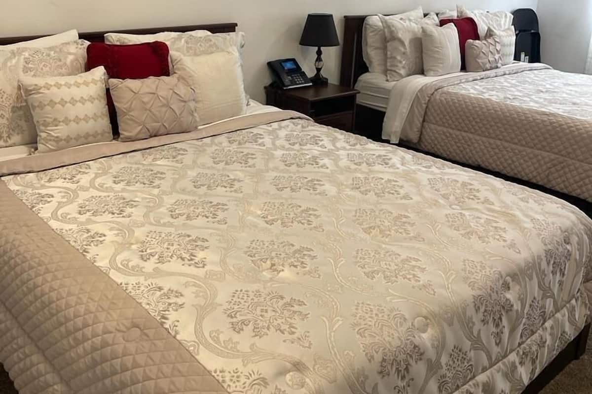 A hotel room with two beds, each with a quilted bedspread and multiple pillows. There is a nightstand with a lamp and a phone between the beds.