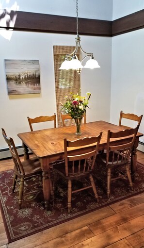 Uma sala de jantar com uma mesa de madeira, seis cadeiras, um vaso com flores, um candeeiro pendente e um quadro na parede.
