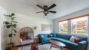 A living room with a blue sectional sofa, a wicker chair, a ceiling fan, and a large window with a view of a garden.