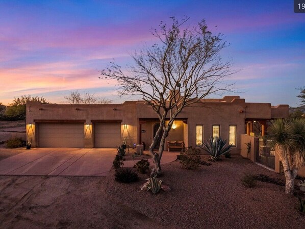 A modern house with a garage, a well-maintained garden, and a clear sky at dusk. - Serene Scottsdale Home:Heated Pool,HotTub,FirePit,Putting Green & Mountain Views (Scottsdale)