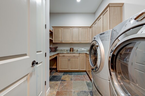 A laundry room with a front-loading washing machine, beige cabinets, a tiled floor, and a countertop with a basket and towels. - Serene Scottsdale Home:Heated Pool,HotTub,FirePit,Putting Green & Mountain Views (Scottsdale)