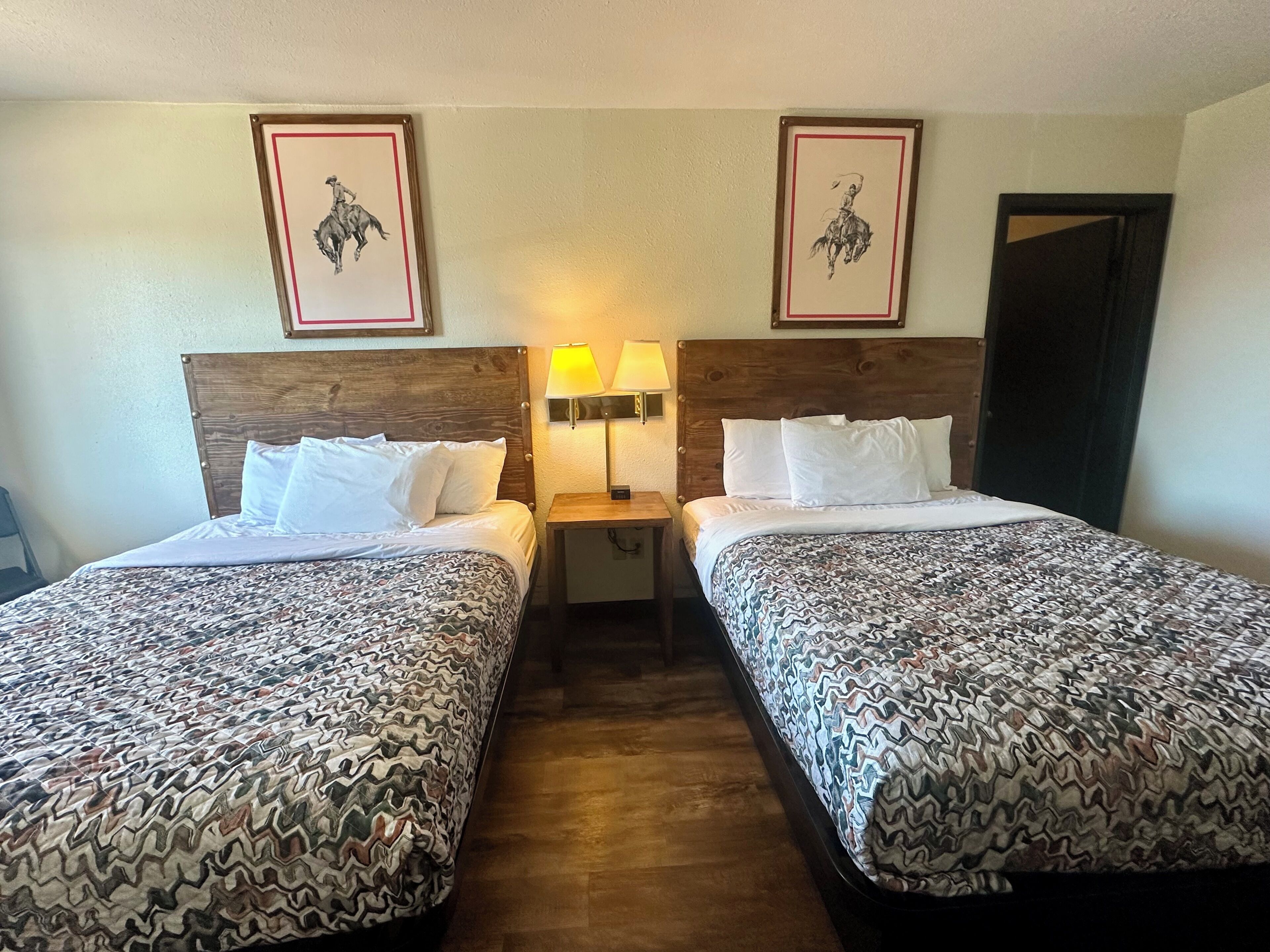 A hotel room with two beds, each with patterned bedding, a wooden headboard, a nightstand with a lamp, and two framed pictures on the wall.