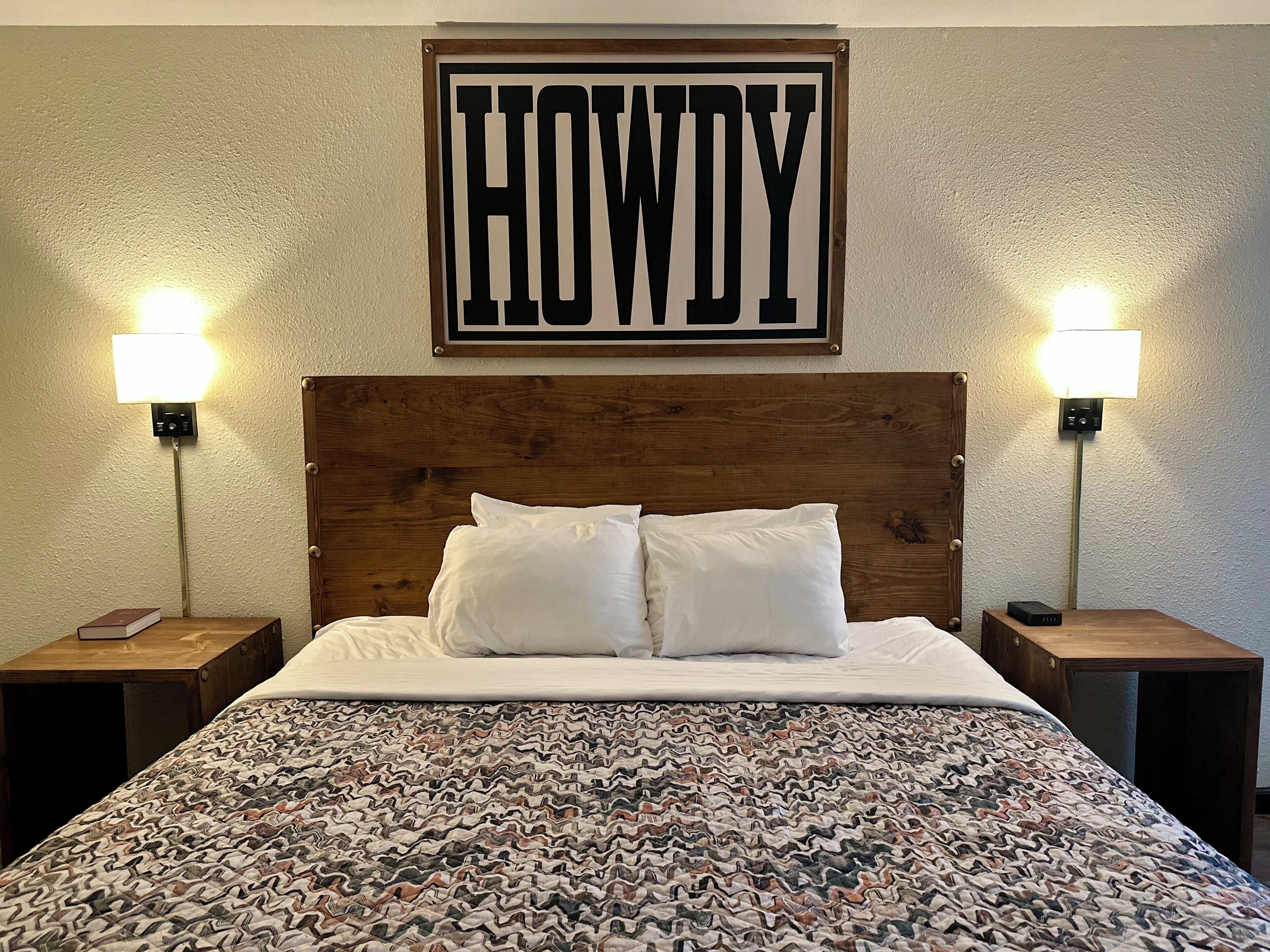 Una cama con una colcha estampada, dos lámparas de noche, un cabecero de madera y un letrero sobre la cama que dice 'HOWDY'.