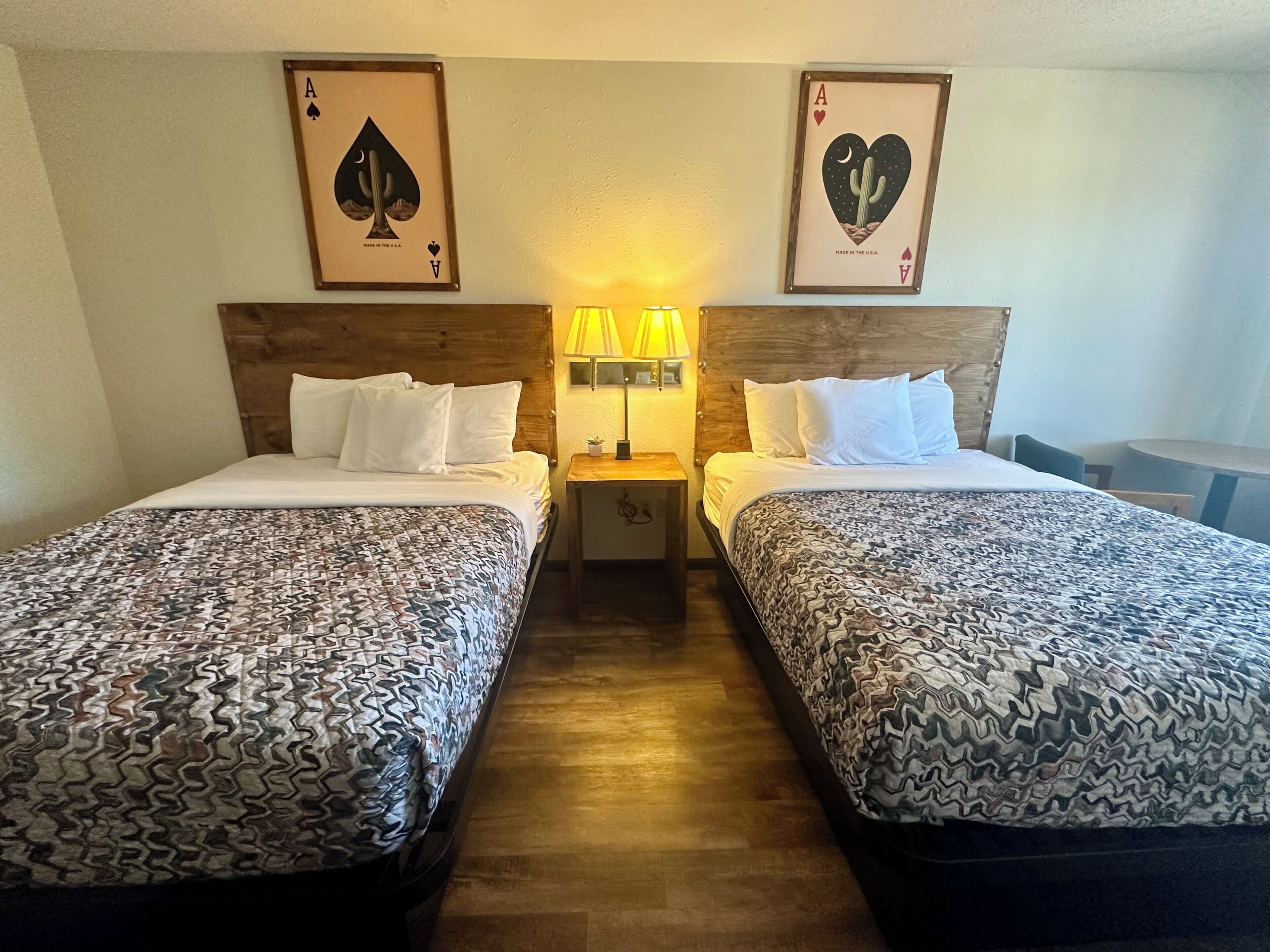 A hotel room with two beds, each with patterned bedding, a nightstand with a lamp, and two framed playing card pictures on the wall.