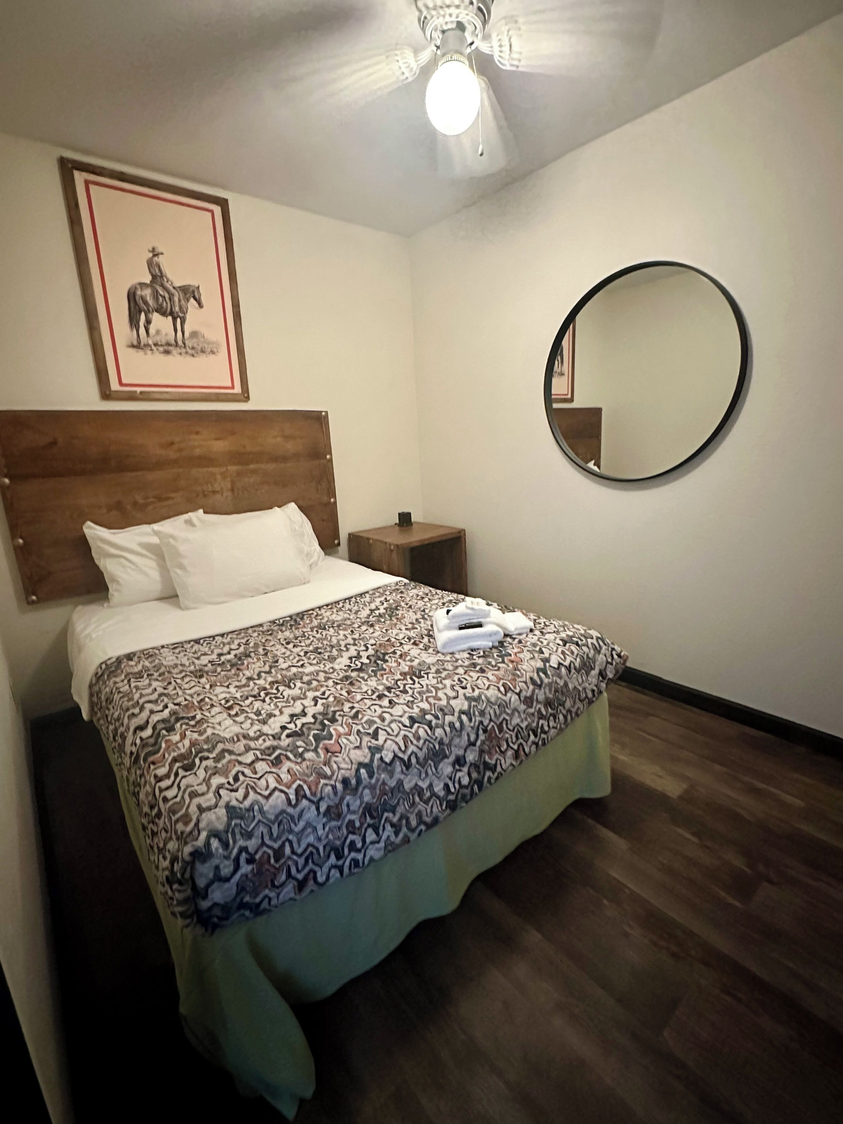 Un dormitorio con una cama, cabecera de madera, mesita de noche, espejo redondo y ventilador de techo.