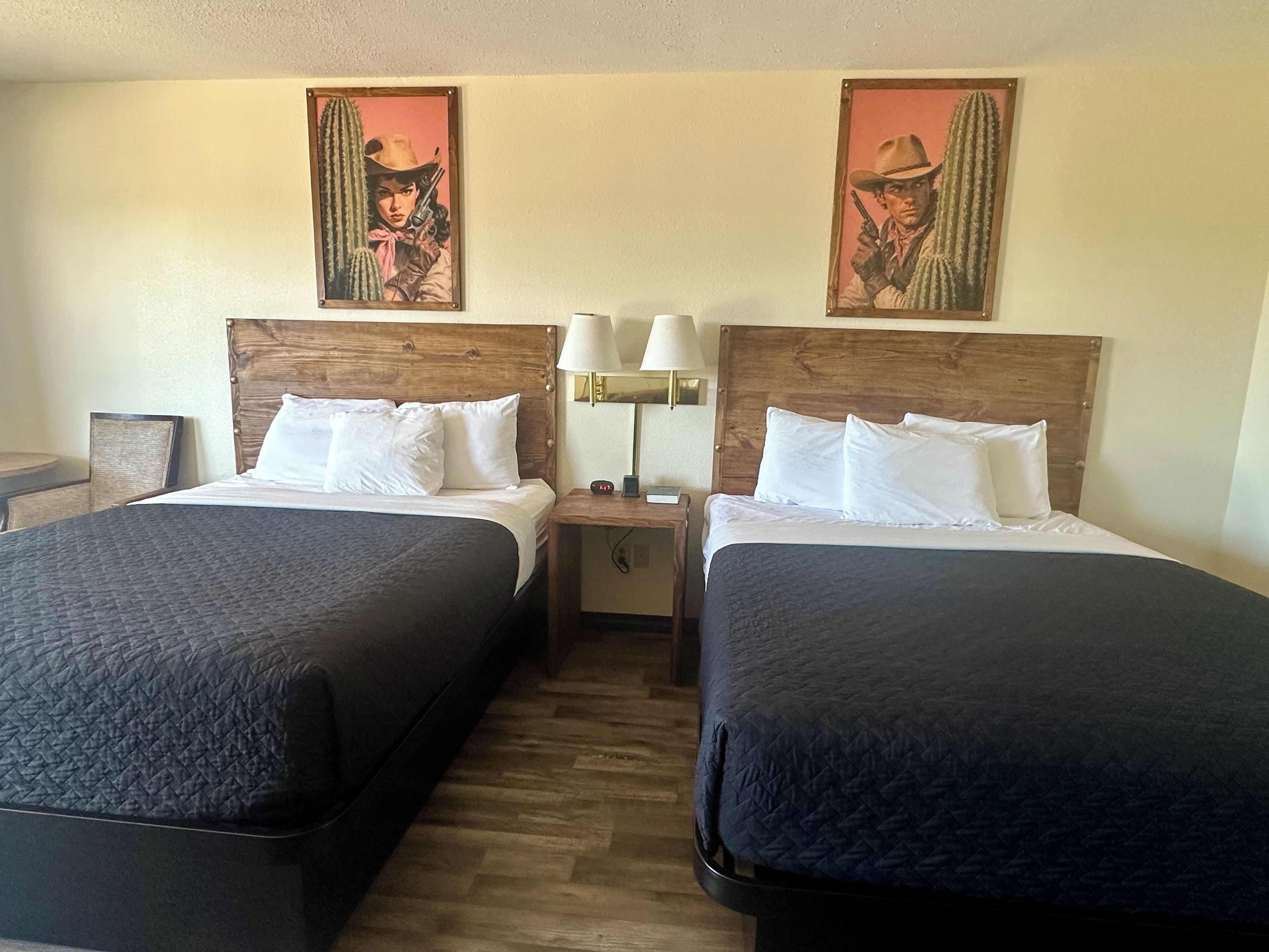 A hotel room with two beds, wooden headboards, white bedding, a nightstand with a lamp, and two wall-mounted paintings.