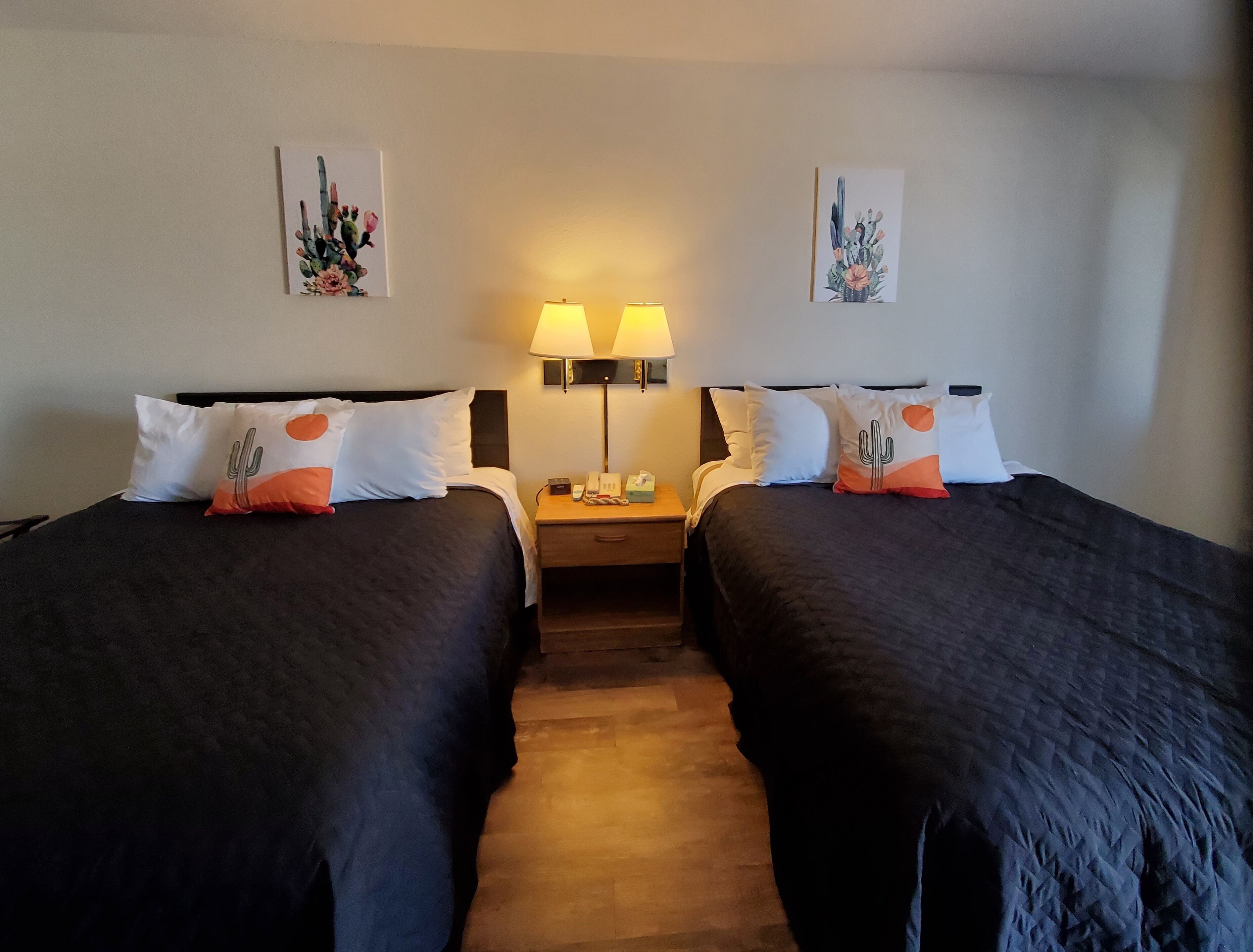 Une chambre d’hôtel avec deux lits, chacun agrémenté de draps foncés et d’oreillers à motifs de cactus. On y trouve deux lampes de chevet et une table de chevet avec un téléphone et une boîte de mouchoirs. La chambre a un plancher en bois et des murs blancs, le plafond étant décoré d’œuvres d’art sur le thème des cactus.