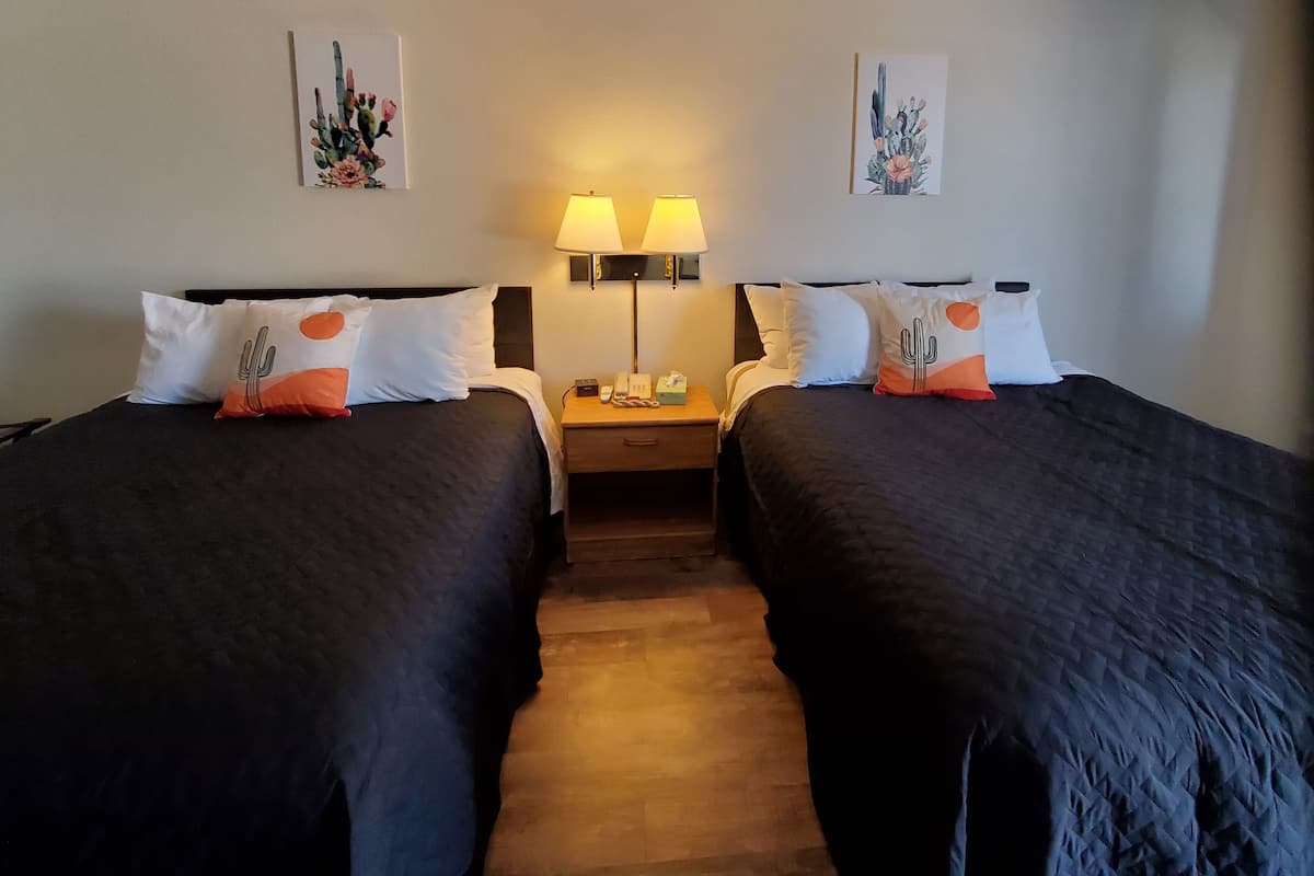 Une chambre d’hôtel avec deux lits, chacun agrémenté de draps foncés et d’oreillers à motifs de cactus. On y trouve deux lampes de chevet et une table de chevet avec un téléphone et une boîte de mouchoirs. La chambre a un plancher en bois et des murs blancs, le plafond étant décoré d’œuvres d’art sur le thème des cactus.