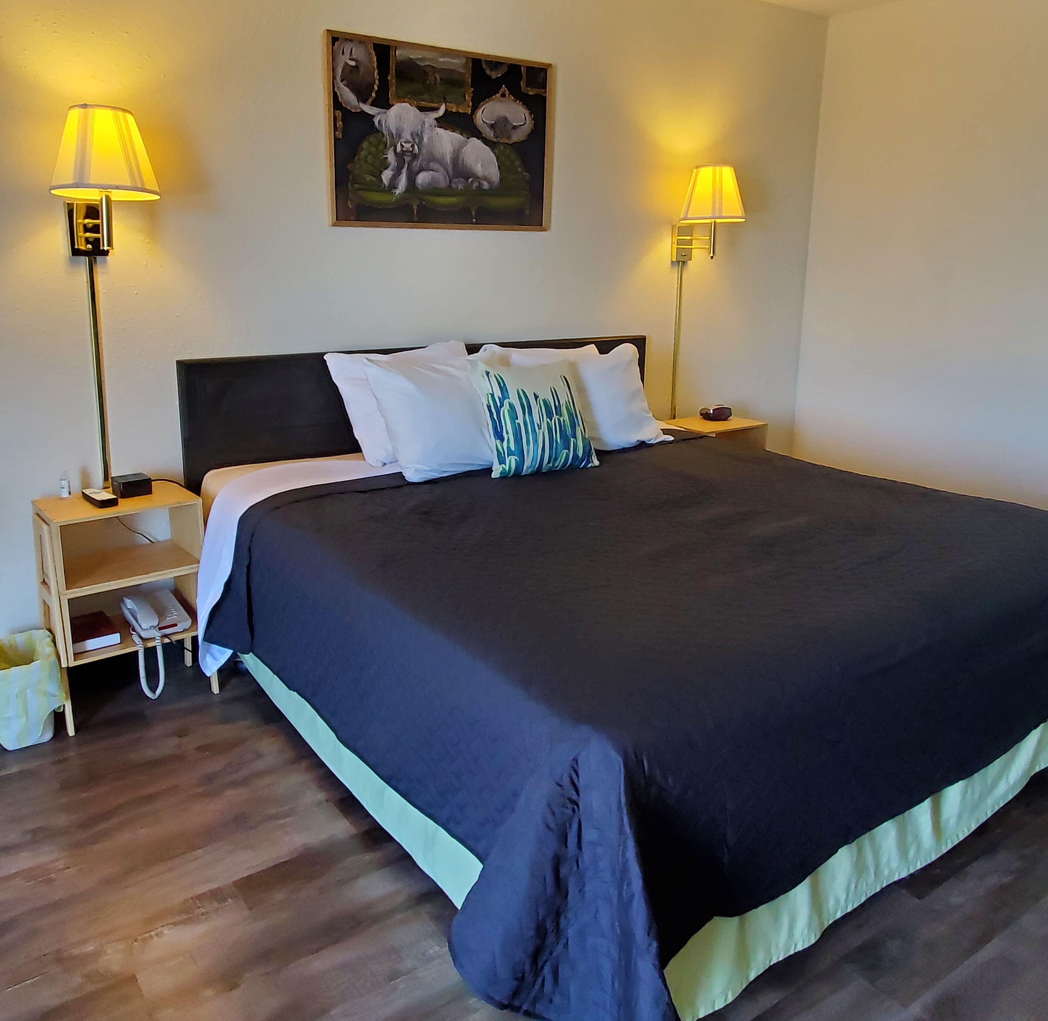 A neatly made bed with a dark-colored duvet, two bedside lamps, a nightstand with a phone and a small item, and a painting above the bed.