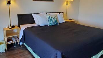A neatly made bed with a dark-colored duvet, two bedside lamps, a nightstand with a phone and a small item, and a painting above the bed.
