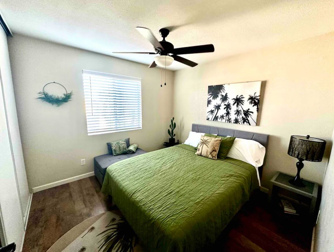 A bedroom with a bed, a ceiling fan, a window with blinds, a wall art piece, and a nightstand with a lamp.
