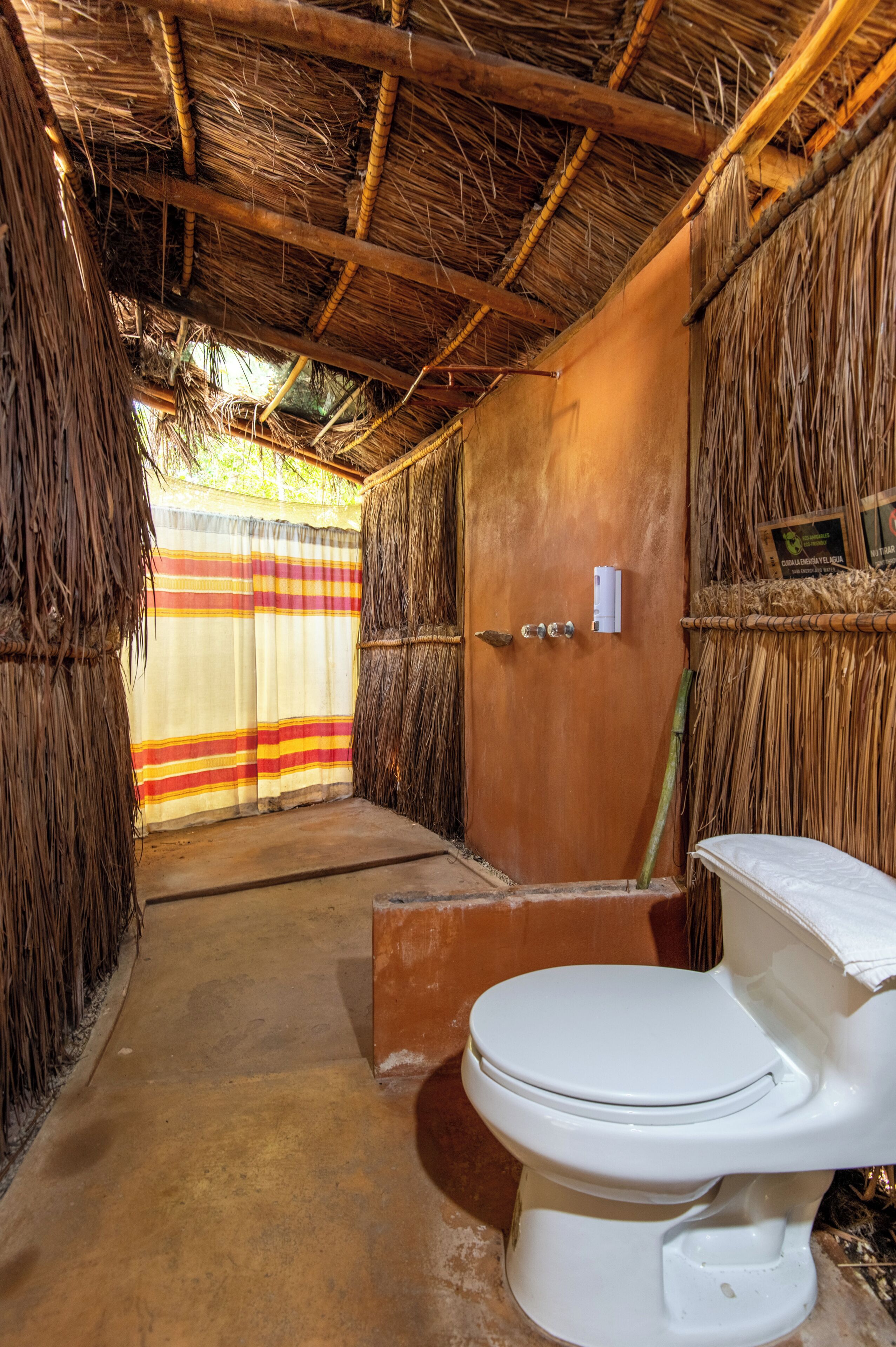 A rustic bathroom with a thatched roof, a striped curtain, and a white toilet.