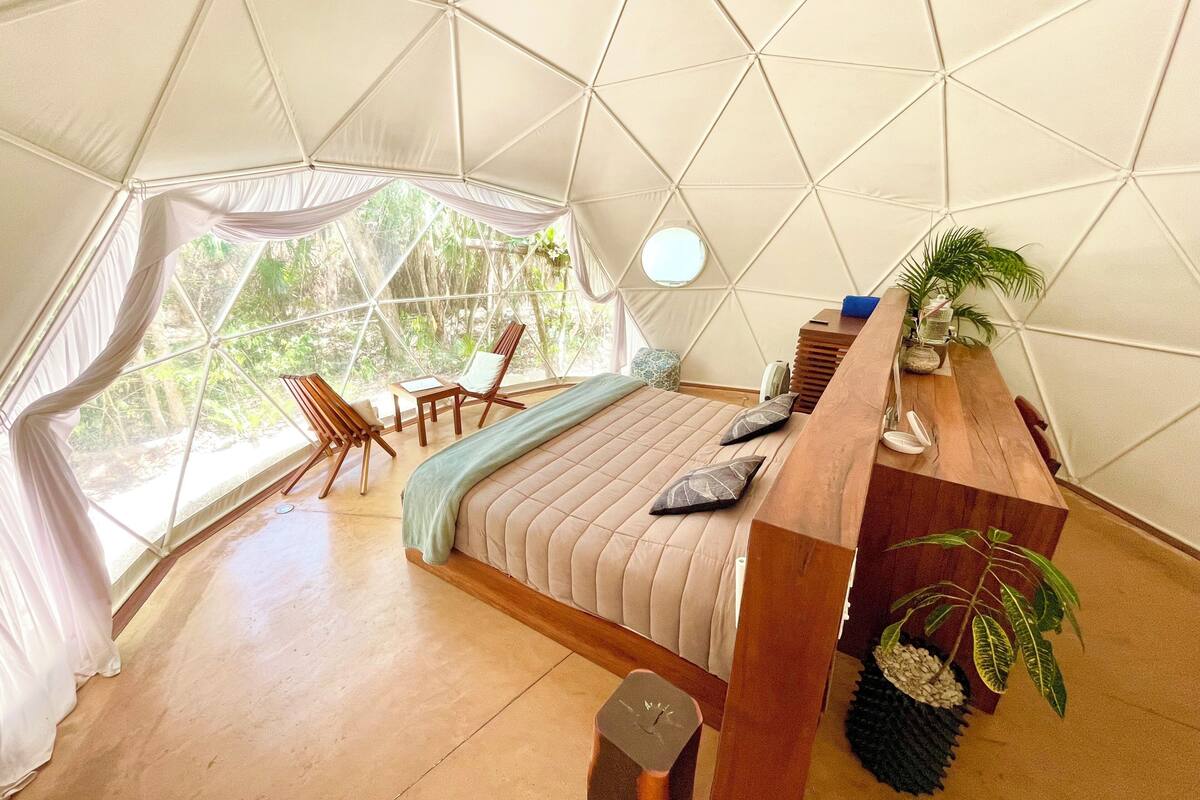 Een tentkamer met een bed, een houten nachtkastje, stoelen en uitzicht op groen buiten.