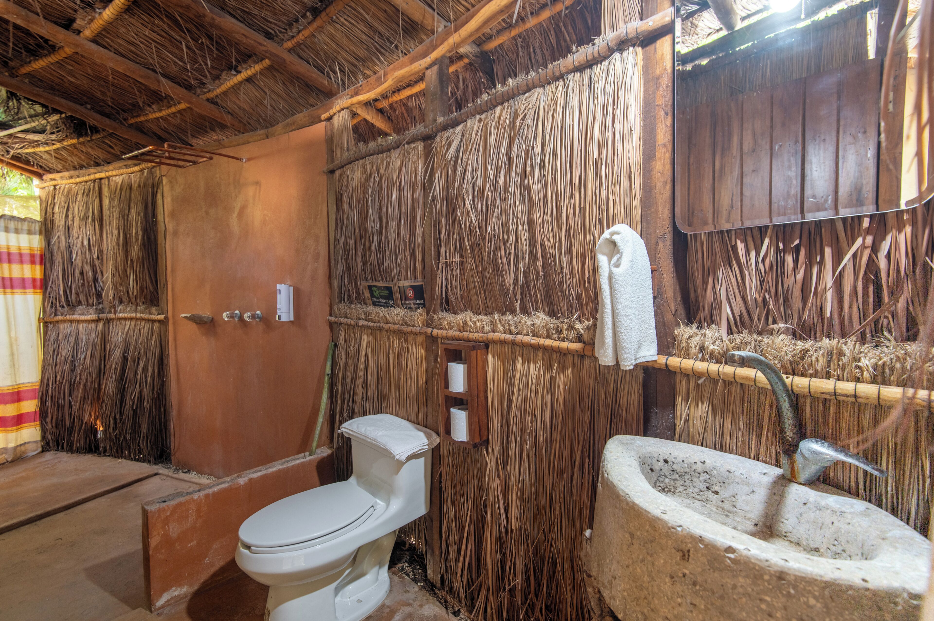 A rustic bathroom with a thatched roof, a white toilet, and a concrete sink.