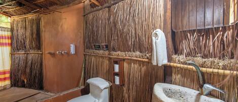 A rustic bathroom with a thatched roof, a white toilet, and a concrete sink.