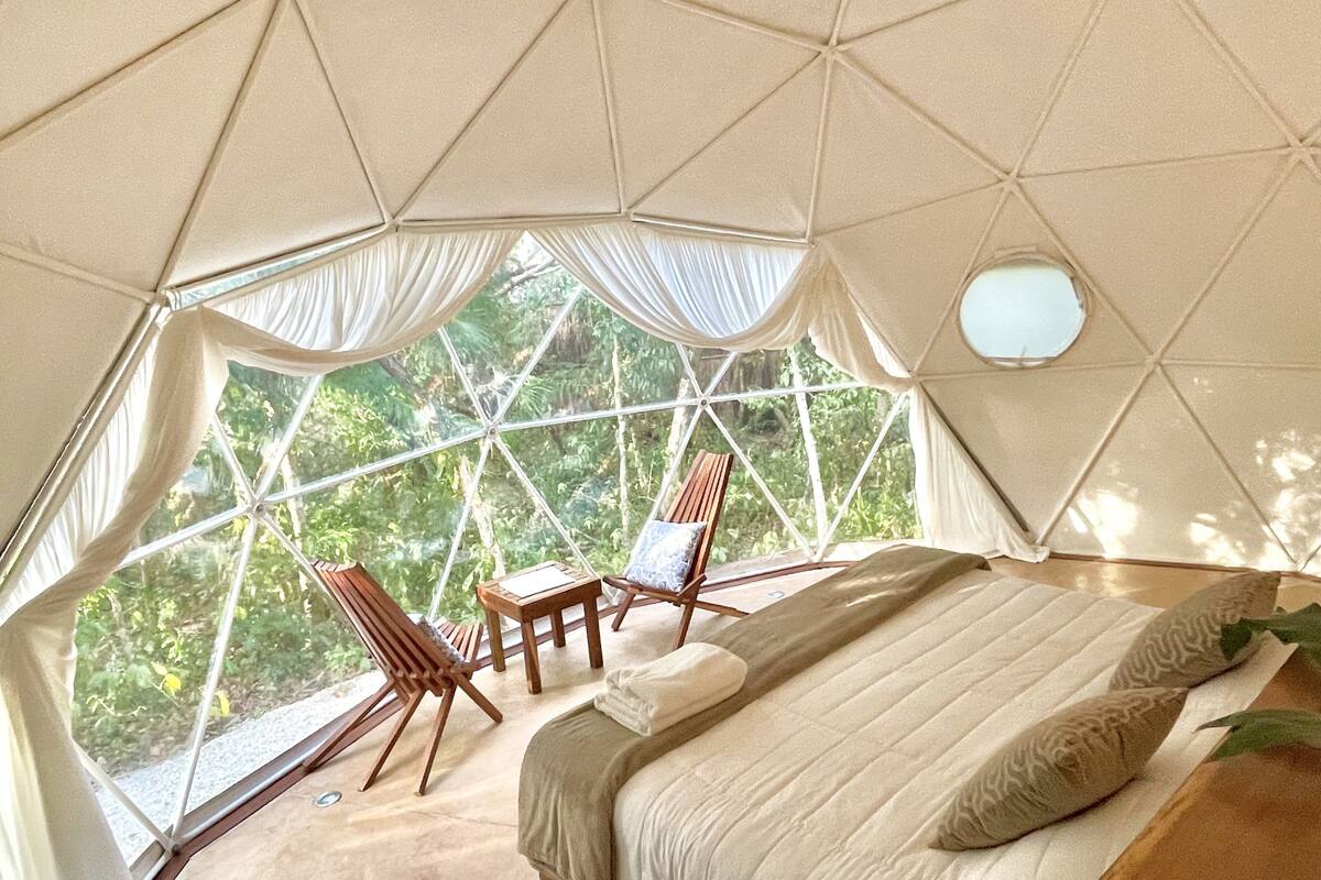 Een kamer in een geodetische koepel met een bed, twee stoelen, een klein tafeltje en een raam.