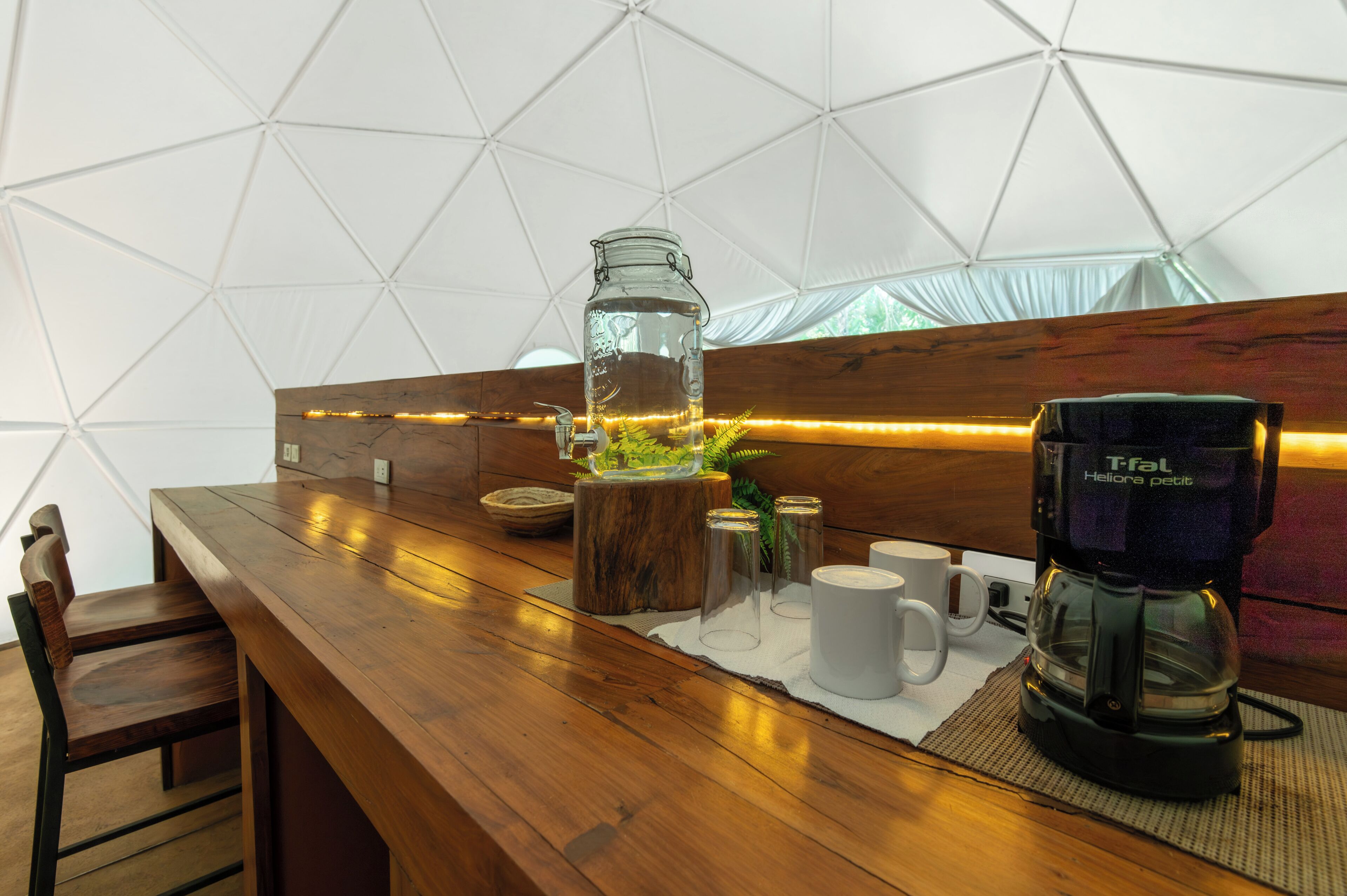 Una mesa de madera con una cafetera, un tarro de vidrio y una planta en una estructura tipo tienda de campaña.