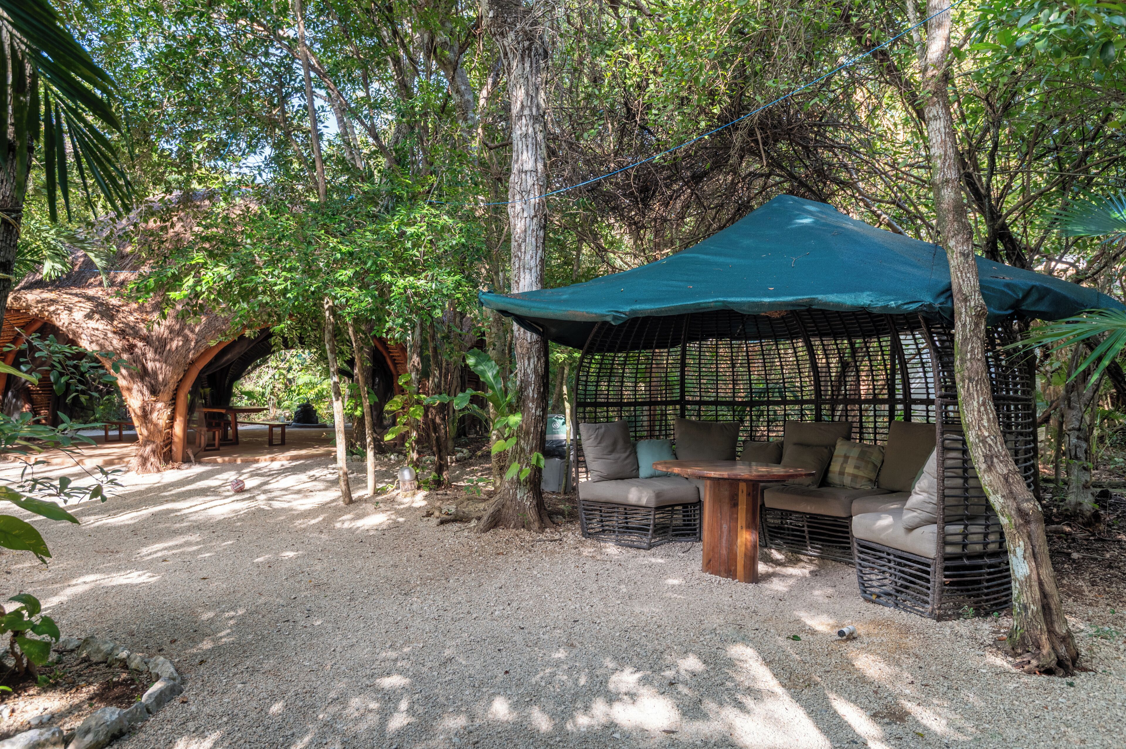 Un espacio de descanso cubierto con mobiliario de mimbre y una mesa, rodeado de árboles y un sendero de grava.