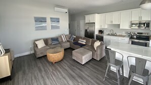 A modern living room with a sectional sofa, a round coffee table, and a kitchen area with white cabinets and stainless steel appliances. - Direct Ocean Front Condo with large wrap around balcony (Hull)