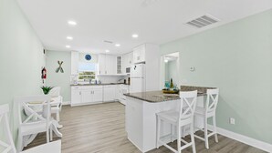 A modern kitchen with white cabinetry, a central island with bar stools, and a dining area with white chairs. - Heart of Flagler Beach-1 Blk 2 Beach-Pets Permitted-Walk EVERYWHERE! (Flagler Beach)