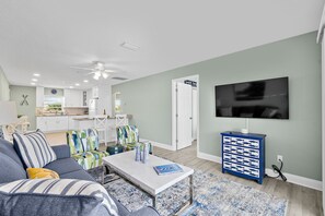 A modern living room with a sofa, coffee table, and a TV. There is a kitchen in the background with white cabinets and a dining area.