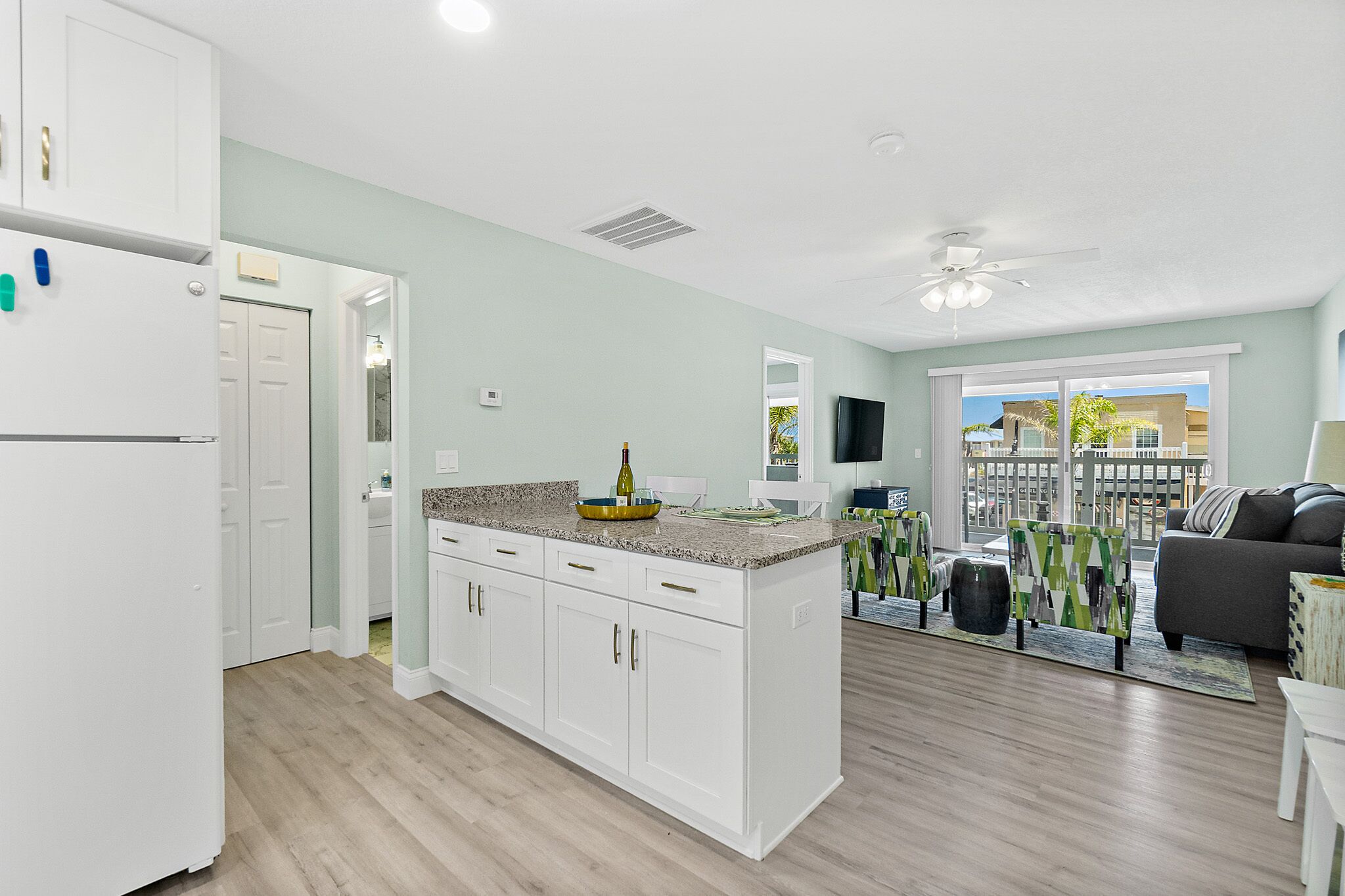 A modern kitchen with white cabinets, a granite countertop, and a built-in island. There is a dining area with a gray sofa and a view of the outside.