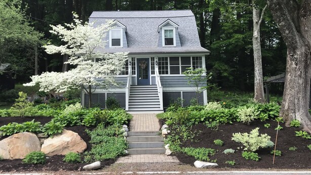 A two-story house with a front porch, surrounded by a garden with various plants and rocks. - Walk into town, soak up sun on enclosed porch & steps away from nature trails! (Stockbridge)