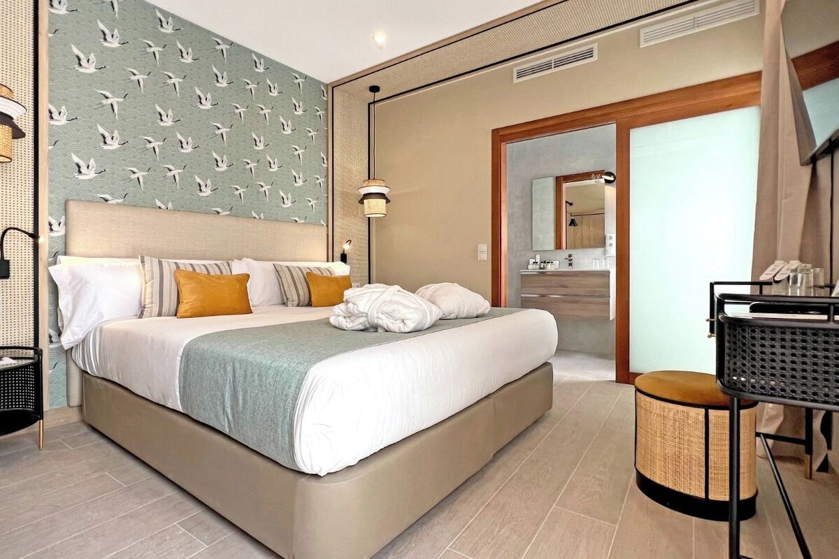 Ett modernt hotellrum med en stor säng, ett sängbord, en spegel och ett badrum som syns genom en öppen dörr.